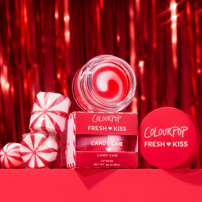 ColourPop Fresh Kiss Candy Cane Lip Mask jar opens to show glossy red balm, stacked with its red 0.28 oz boxes beside peppermint candies, set against a shiny red tinsel holiday backdrop for a festive, gift-ready beauty shot.