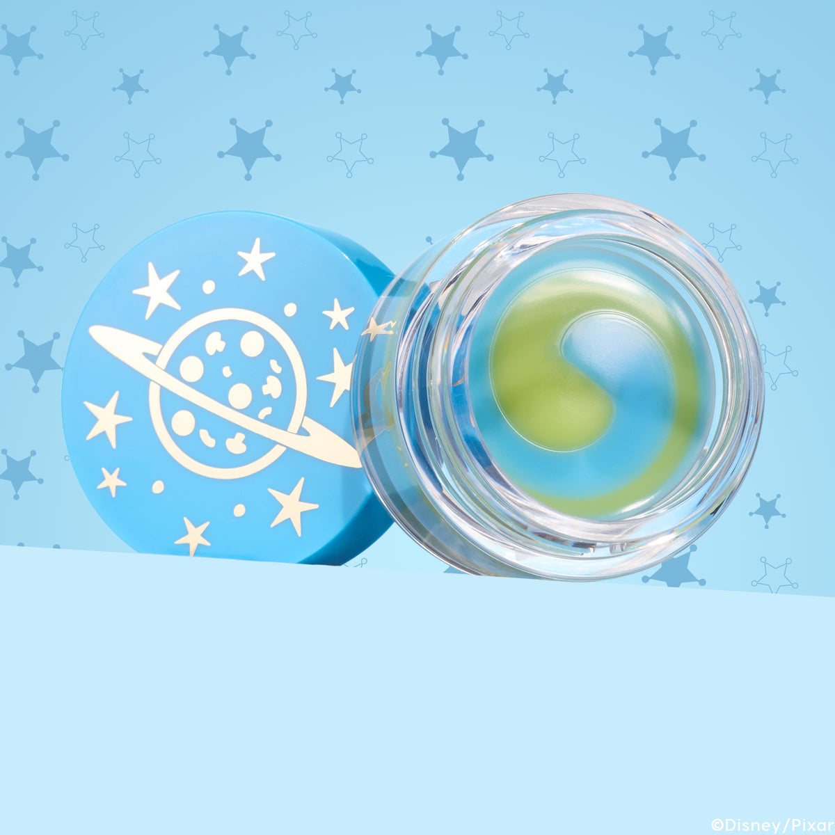 Clear jar of The Chosen One ColourPop Lip Mask showing a yin-yang swirl of green and blue balm, resting open beside a sky-blue lid printed with a ringed planet and stars, set on a light blue background with star-badge patterns.