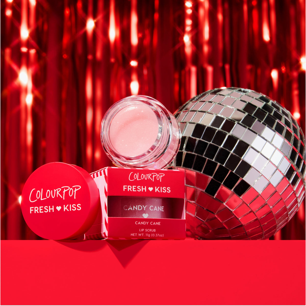 ColourPop Fresh Kiss Candy Cane lip scrub, open to show pink shimmering scrub in a clear jar, posed with its red lid and box, set against glossy red tinsel and a silver disco ball for a festive holiday beauty scene.