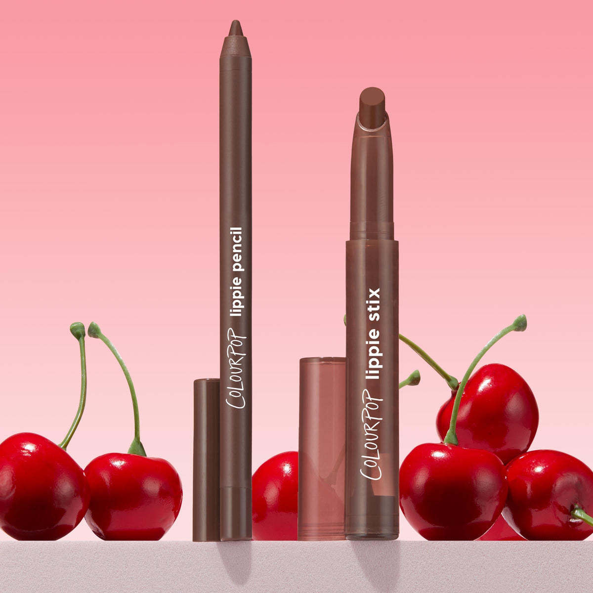 ColourPop Lippie Pencil in Pitch stands upright with cap off beside a matching Lippie Stix, both in a deep brown shade, set among glossy red cherries on a pink gradient backdrop for a bold, cherry-inspired lip look.