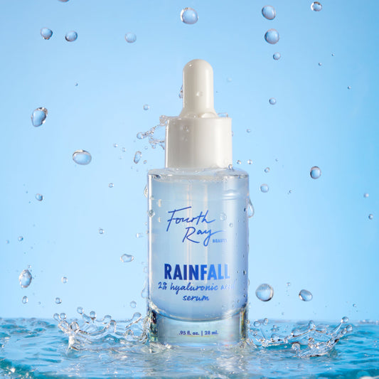 Fourth Ray Beauty Rainfall 2% HA Serum dropper bottle standing upright, catching water splashes against a light-blue backdrop, showcasing its hydrating hyaluronic acid formula; ColourPop Face Serum, 0.95 fl oz/28 mL.