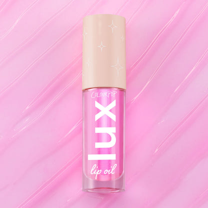 ColourPop Lux Lip Oil in Raspberry Mojito, a clear tube with bold “LUX” lettering and a beige star-print cap, displayed on a glossy swirled pink background, showcasing the vibrant pink-tinted lip oil formula.