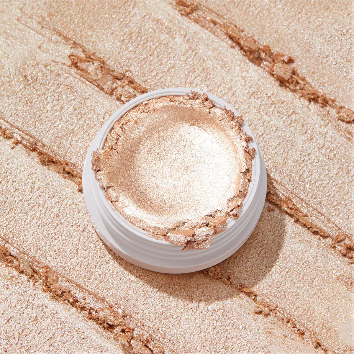 ColourPop Flexitarian Super Shock Highlighter gleaming in an open white pot, its creamy champagne‑pearl formula swatched in broad metallic streaks across the backdrop, showcasing intense shine and a smooth, blendable texture.