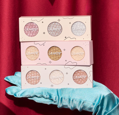 ColourPop Matcha and Mochi Super Shock Shadow trio, three pastel palettes of shimmery eyeshadows stacked on a teal satin glove against a red curtain backdrop, showcasing glittery metallic shades for vibrant eye looks.