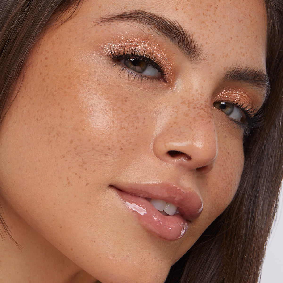 Glossy lips wearing ColourPop So Juicy Plumping Glossy Lip Oil in Lychee Jelly, plumping and high-shine; model tilts face as bronze shimmer eyeshadow and natural freckles catch studio light in a close-up beauty shot.