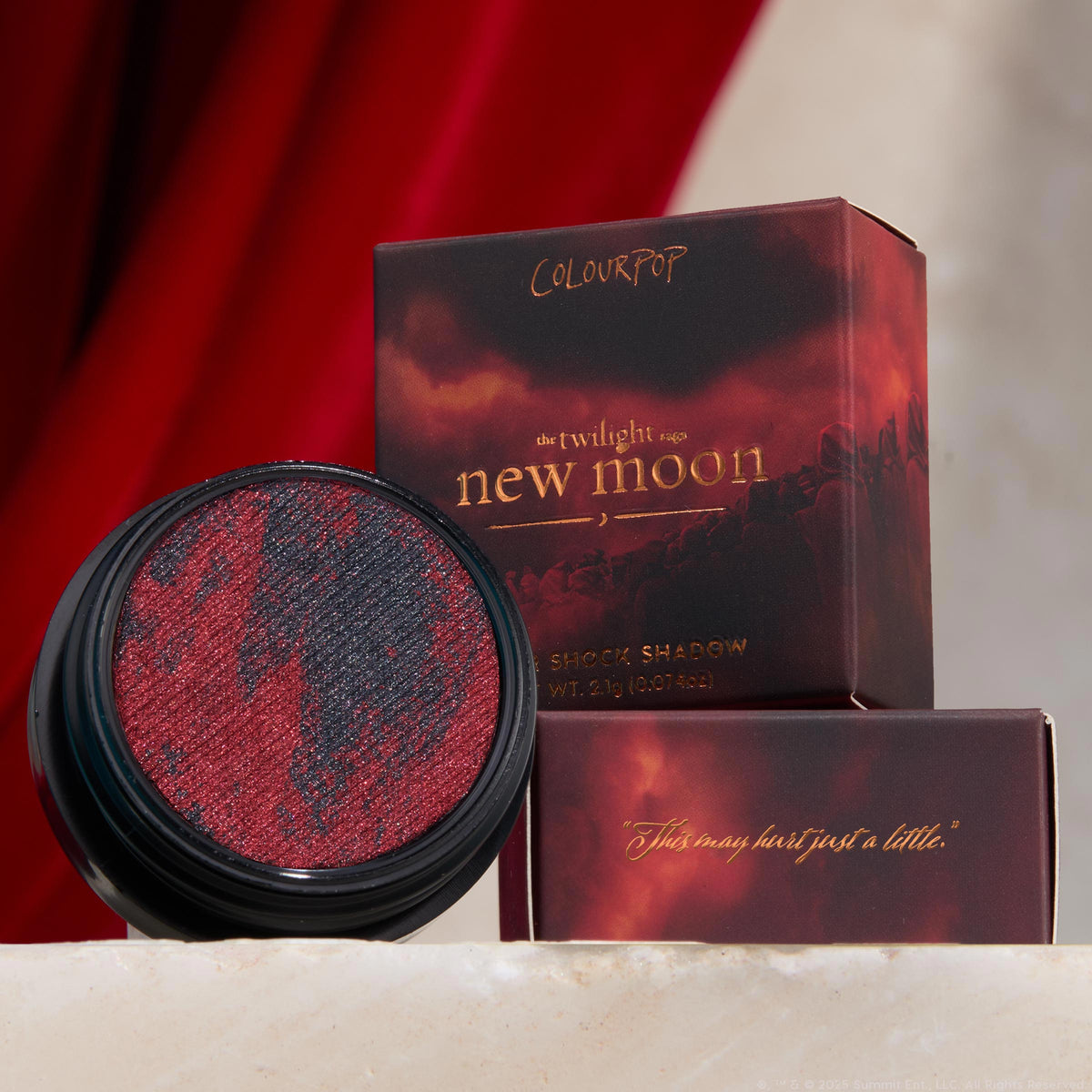 The Volturi ColourPop Super Shock Shadow showcases marbled crimson and charcoal shimmer in a black pot, catching light as it sits before Twilight: New Moon–themed boxes with gold lettering, set on a stone ledge against a rich red velvet backdrop.