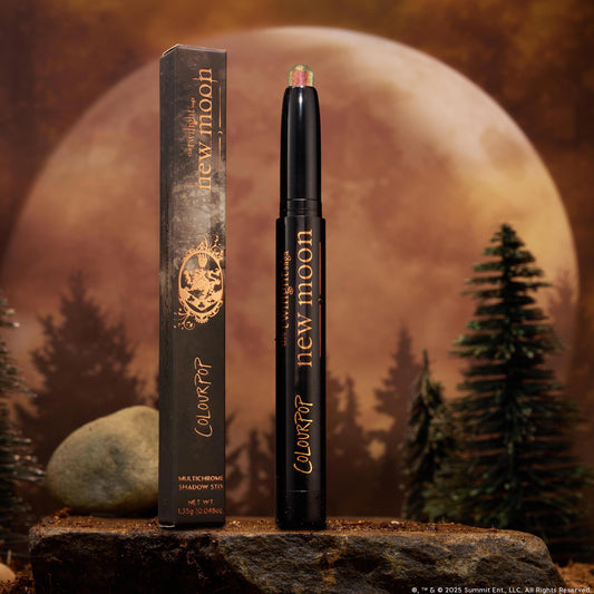 Vampire Girl ColourPop Shadow Stix stands uncapped, showing a pink‑green multichrome tip on a sleek black tube with gold lettering, its box beside it, set on a rocky surface with pine trees and a glowing full moon background.