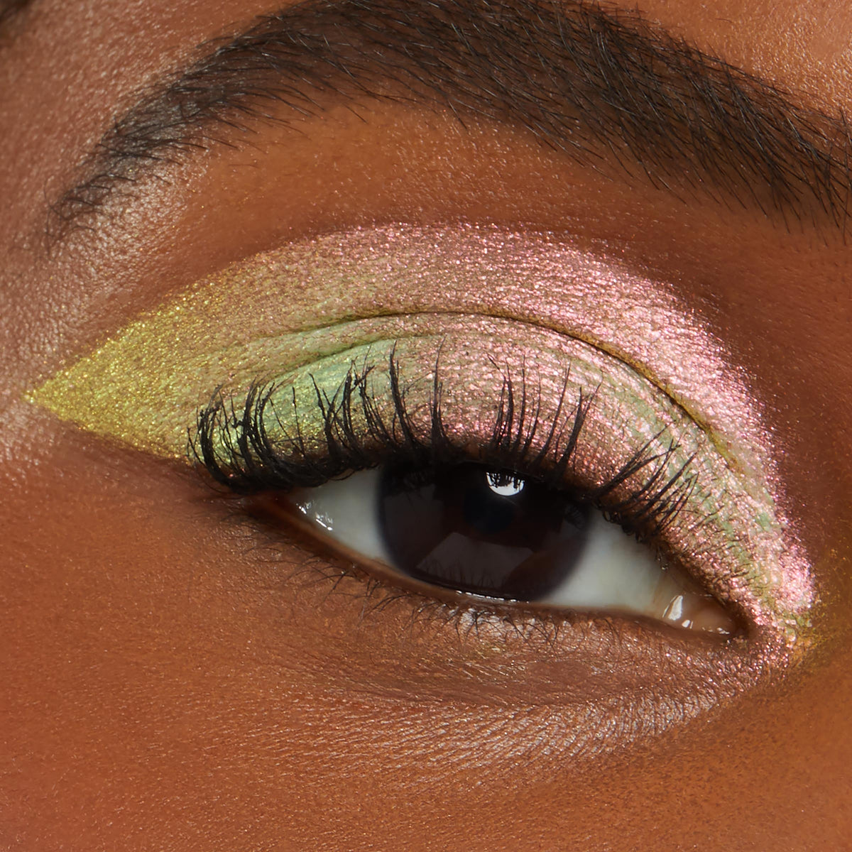 Close-up eye wearing Galaxy Shadow Stix eyeshadow, showing a bold cut-crease look in metallic pink, green, and gold shimmer on deep skin, highlighting the multi-chrome pigment and smooth, blendable finish for dramatic glam makeup.