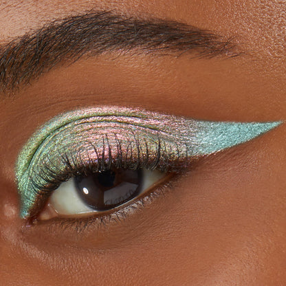 Close-up of an eye wearing Colourpop Game Over Shadow Stix, showcasing a sharp metallic teal wing blended into multicolor iridescent pink and green shimmer across the lid on smooth medium-brown skin.
