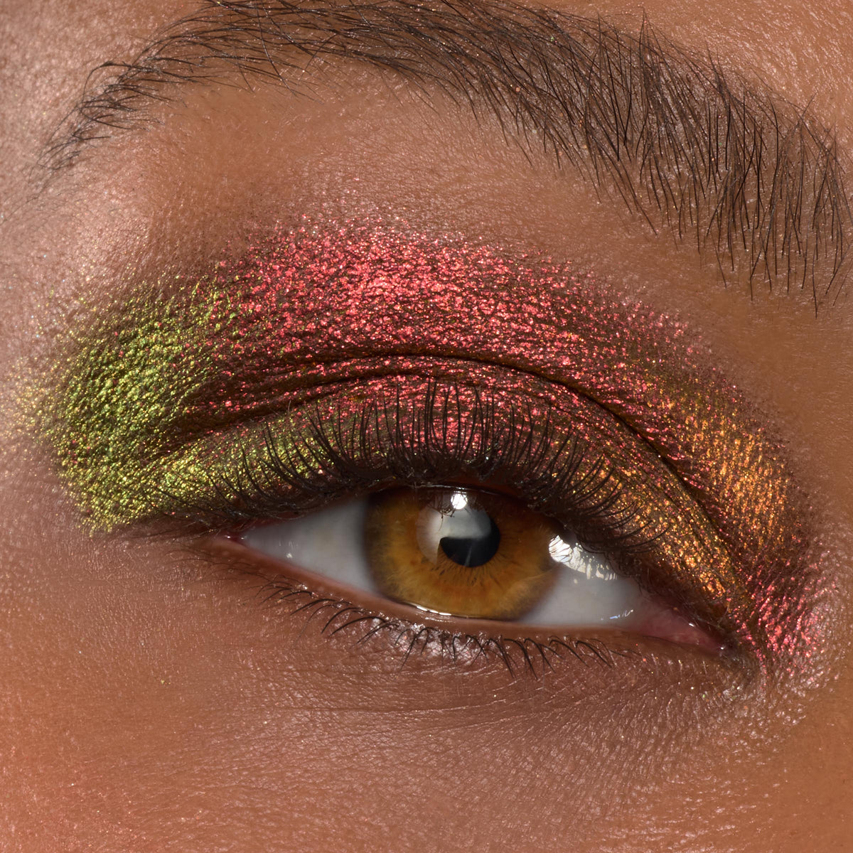Shimmering multichrome eyeshadow illuminates a hazel eye, with red-rose, copper, and lime-green shifts swept across the lid using ColourPop Shadow Stix in Love Triangle, showcasing bold pigment and a metallic finish in a close-up.