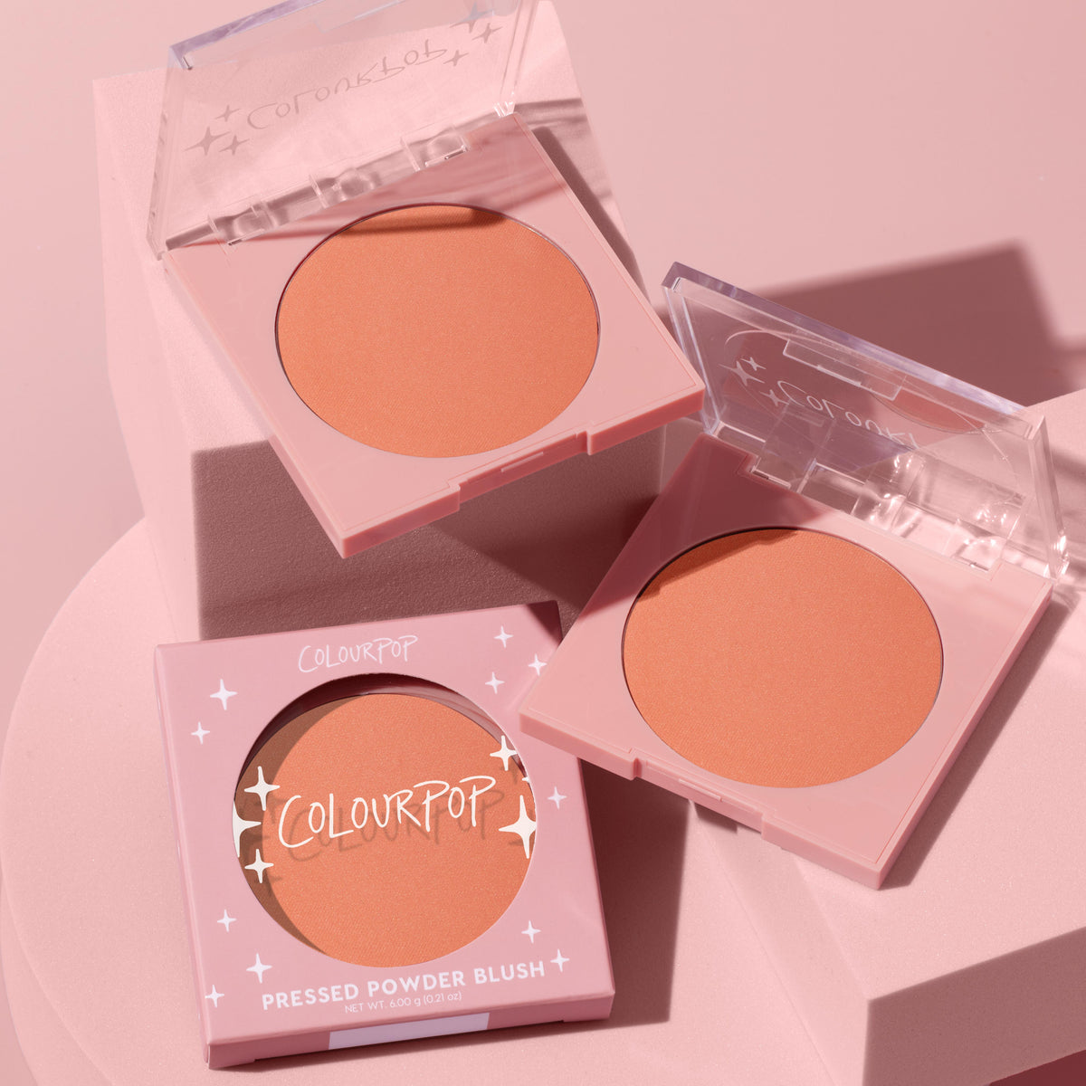 ColourPop Pressed Powder Blush Shortbread compact displays a warm peach matte pan in a pale pink square case, with one boxed and two open compacts arranged on a soft pink pedestal under studio lighting.