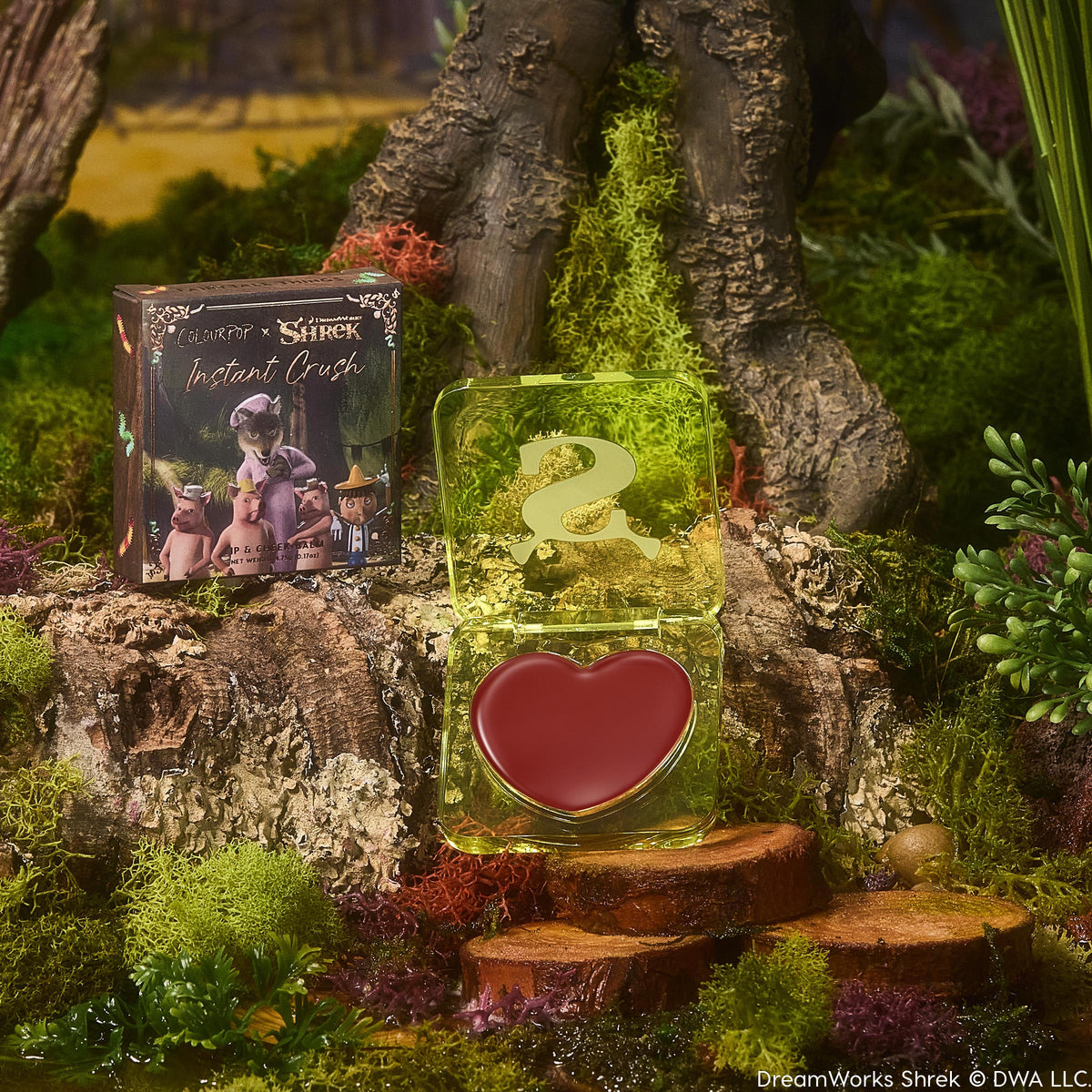 Fairytale Things ColourPop Lip and Cheek Balm opens to reveal a heart-shaped rosy tint in a translucent lime-green compact with an S logo, displayed on a mossy tree stump beside the Shrek collab carton in a whimsical swamp scene.