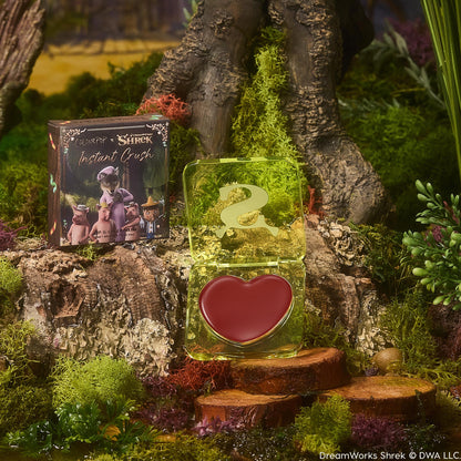 Fairytale Things ColourPop Lip and Cheek Balm opens to reveal a heart-shaped rosy tint in a translucent lime-green compact with an S logo, displayed on a mossy tree stump beside the Shrek collab carton in a whimsical swamp scene.