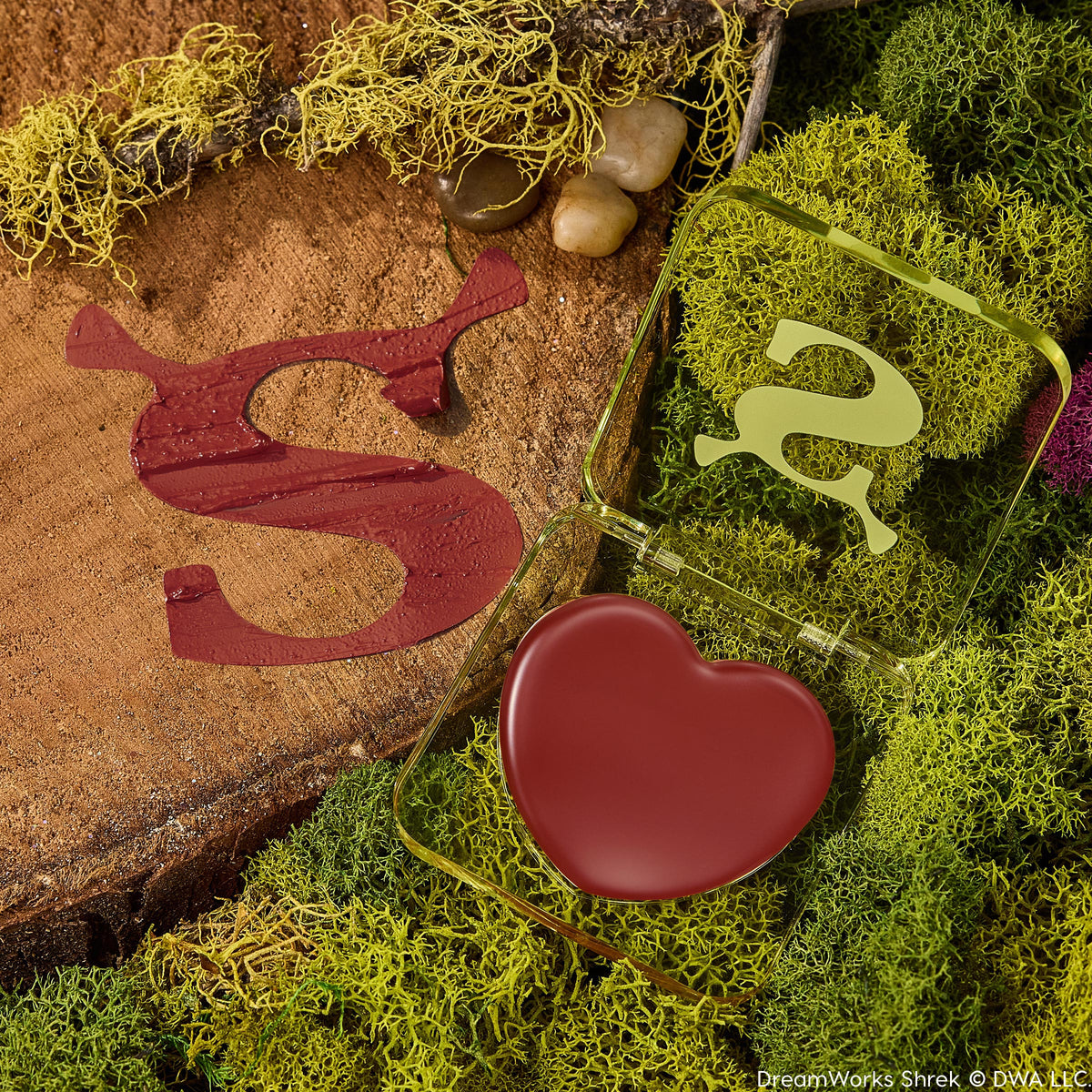 Fairytale Things ColourPop Lip and Cheek Balm sits open, revealing a heart-shaped pan of warm brick-red pigment in a translucent green, Shrek-inspired compact with an ogre-ear S, resting on moss and a wood slice beside a glossy burgundy S swatch.