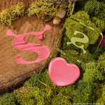 Heart-shaped pink balm in a translucent green compact sits open on moss and a wood slab with a Shrek-style S, showcasing ColourPop Lip and Cheek Balm, True Love’s Form, a lip and cheek tint in a playful swamp-inspired setting.