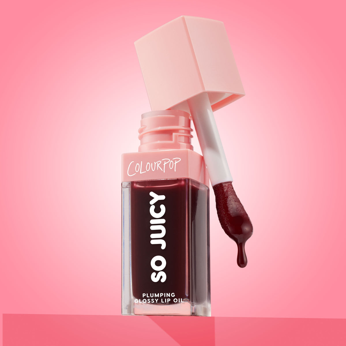 ColourPop So Juicy Plumping Glossy Lip Oil in Beverly Drive stands open with the applicator lifted and a drop of deep cherry oil forming, the square clear tube and blush-pink cap set against a glossy pink gradient background.