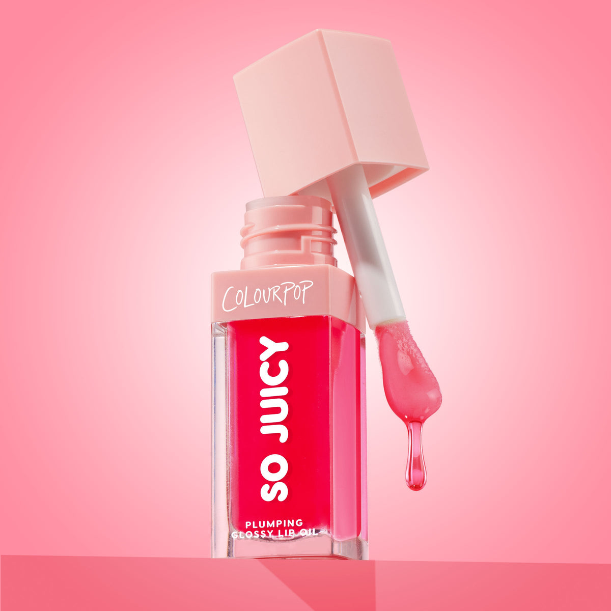 ColourPop So Juicy Plumping Glossy Lip Oil in Flirty Text opens with the doe-foot applicator dripping rosy pink gloss, highlighting the clear square bottle, blush cap, and bold So Juicy logo against a pink gradient studio backdrop.