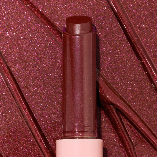 Gloss balm stick twisted up from a pink tube, L-O-V-E shade of the ColourPop So Juicy Plumping Gloss Balm with Peptides, shown close-up against glossy rose-plum shimmer swatches to highlight its dewy, plumping shine and sparkly finish.