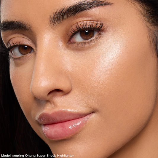 Model wearing Ohana ColourPop Super Shock Highlighter catching light on the cheekbone, delivering a pearly champagne glow with a smooth, dewy finish; close-up beauty shot shows natural skin texture, soft lighting, glossy lips, and defined brows.