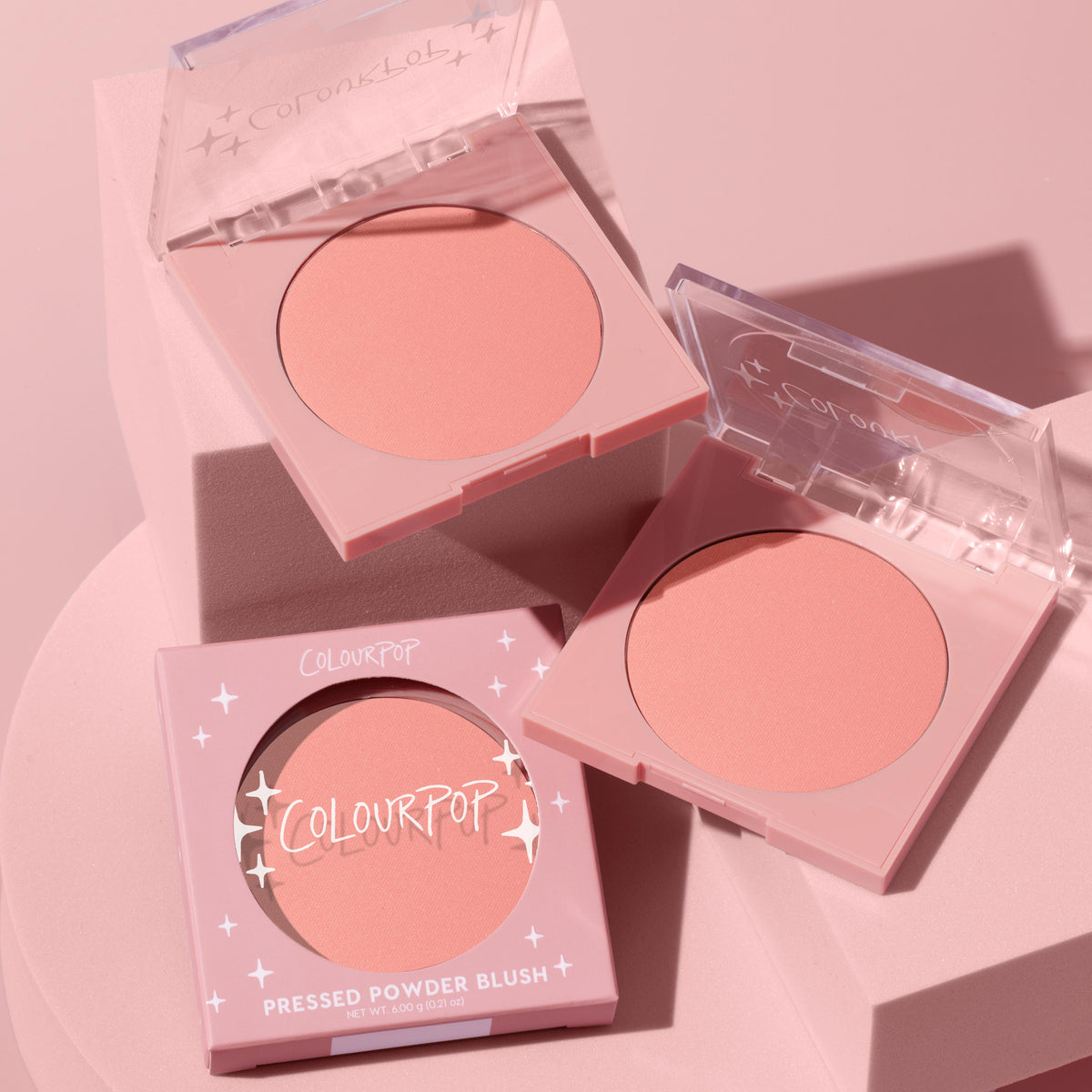 Swirled ColourPop Pressed Powder Blush shown in pastel pink square compacts - two open to reveal a matte peachy‑pink pan, one in branded box - arranged on blush‑pink blocks under soft studio light for a clean, modern look.