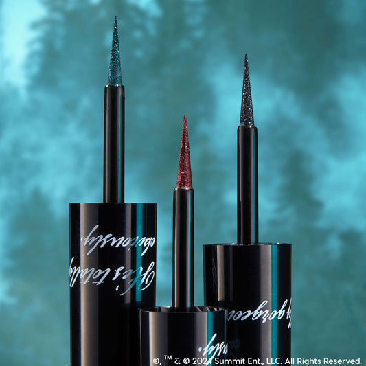 ColourPop Graphix Ink Liner, I Know What You Are, stands uncapped with an ultra-fine glitter brush tip displayed, flanked by two shimmer liners in black tubes with silver script against a teal, forest-blurred backdrop.