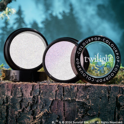 Meadow ColourPop Super Shock Highlighter compact opens to reveal an iridescent silver‑lilac pan, with a Twilight‑branded lid propped beside it, styled on a mossy tree stump against a misty forest backdrop.