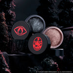 Coven Clash Jelly Much Duo eyeshadows sit open on black fabric, revealing shimmery charcoal and champagne shades in round pots with bold red Twilight-inspired symbols, styled against a dark, moody forest backdrop.