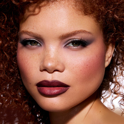 Model wearing ColourPop New Love Lippie Stix from the Twilight collection, showing rich matte deep berry lipstick on full lips with soft-focus glam eye makeup in a close-up beauty shot against a clean studio background.