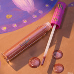 Disney Tangled x ColourPop Ultra Glossy Lip Flynn Rider lies open with doe-foot applicator, revealing shimmery rose-gold gloss in a clear tube; swirled droplets shine on a tan backdrop beside a purple lantern motif and pink cap with iridescent prints.