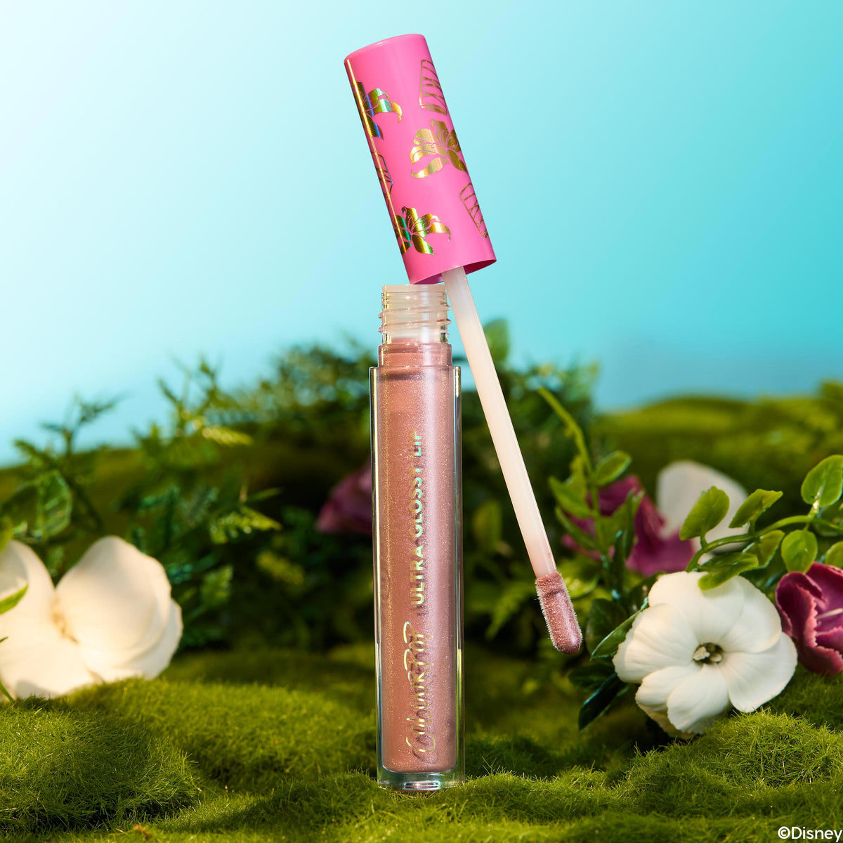 ColourPop Ultra Glossy Lip in Flynn Rider, a shimmery rose-gold gloss, stands open with its doe-foot applicator lifted from the clear tube and pink cap with gold motifs, set on moss with white and purple flowers against a sky-blue backdrop.