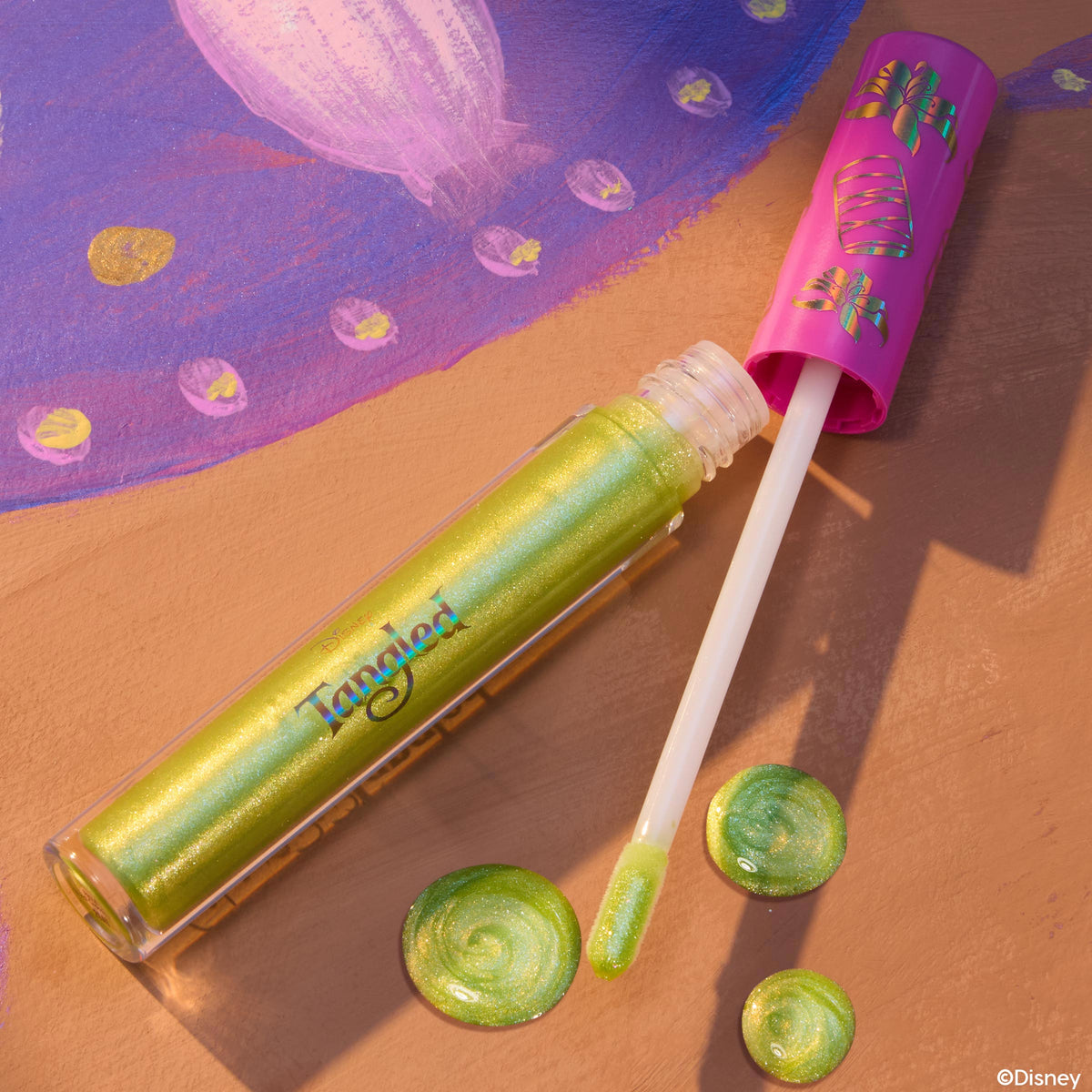 Pascal ColourPop Ultra Glossy Lip lying open with a doe-foot applicator and swirls of lime-green, gold-shimmer gloss, set on a Tangled-inspired painted backdrop, clear tube showing iridescent sheen and pink cap decorated with chameleon icons.