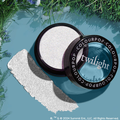 Vampire Skin ColourPop Super Shock Highlighter gleams in an open jar, showing an icy white, sparkly pan with the Twilight-branded lid and a bold swatch, styled as a flat lay on a teal backdrop bordered with green foliage.