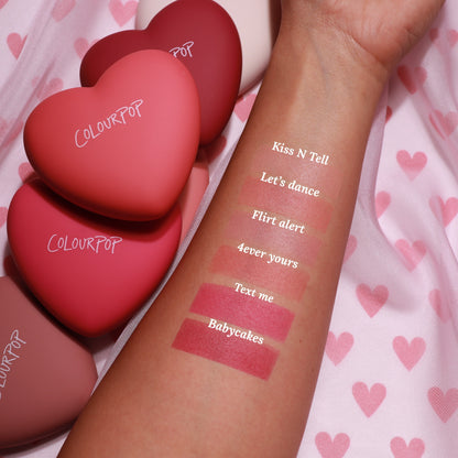 Babycakes pressed powder blush swatch - deep rosy mauve matte - applied on a forearm beneath five labeled shades, with heart-shaped ColourPop compacts on pink heart fabric, highlighting the rich payoff of ColourPop Pressed Powder Blush.