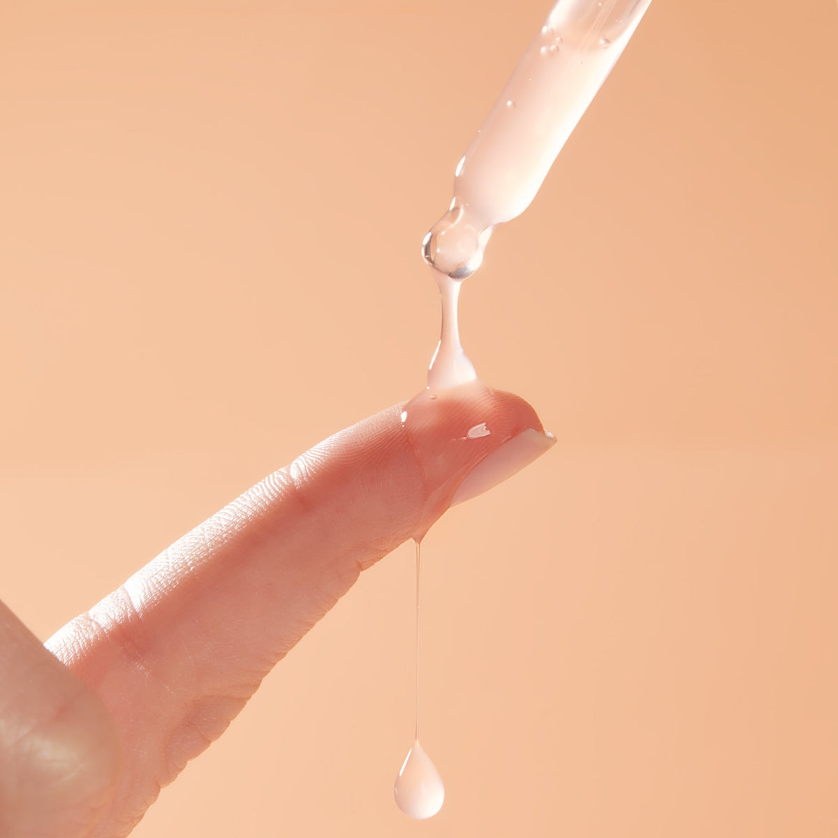 Dropper dispensing milky Reveal 10% AHA Serum onto a fingertip, a glossy bead stretching and dripping against a soft peach background, showcasing the exfoliating ColourPop Face Serum for smooth, radiant skin.