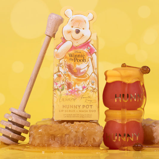 ColourPop Hunny Pot lip care kit features a lip scrub and mask duo, with two honey-colored HUNNY jars stacked and dripping beside a wooden honey dipper, honeycomb, and Disney Winnie the Pooh box art on a bright yellow background.